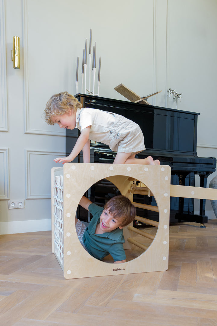 Two children play together on the Babiem Ben Climbing Cube, one climbing on top while the other crawls through the round opening of the natural wooden play cube.