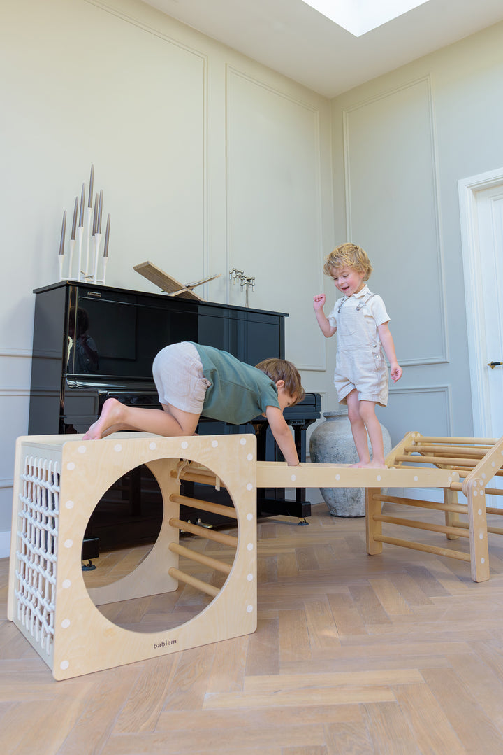 Two children climb and balance on the Babiem Ben Climbing House, exploring the natural wooden indoor play structure with connecting bridge and cube.