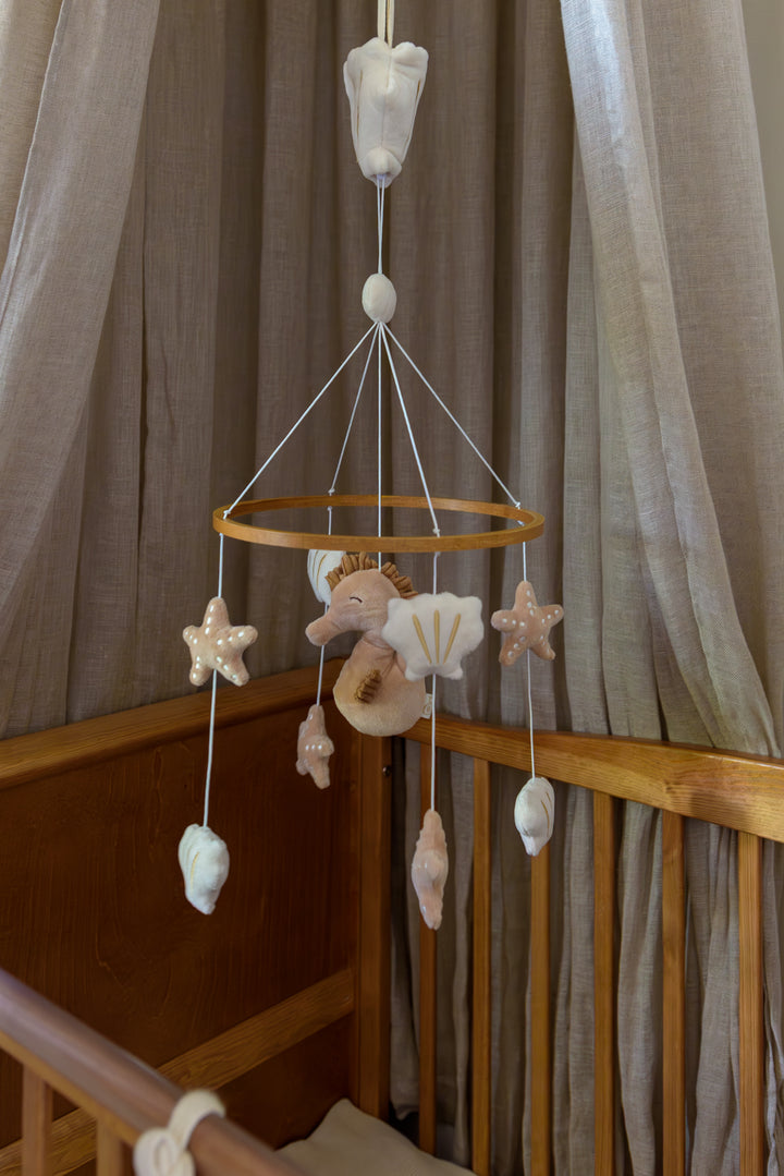A close-up of the Babiem Musical Mobile Zoë hanging above a wooden crib, featuring plush stars, seashells, and a seahorse in soft beige and white tones, creating a soothing and elegant nursery atmosphere.