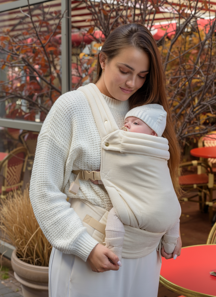 A woman holding her baby in the Babiem baby carrier Olivia while standing outside a café. The soft off-white fabric and cozy design provide warmth and comfort, reflecting Babiem’s calm and natural aesthetic.