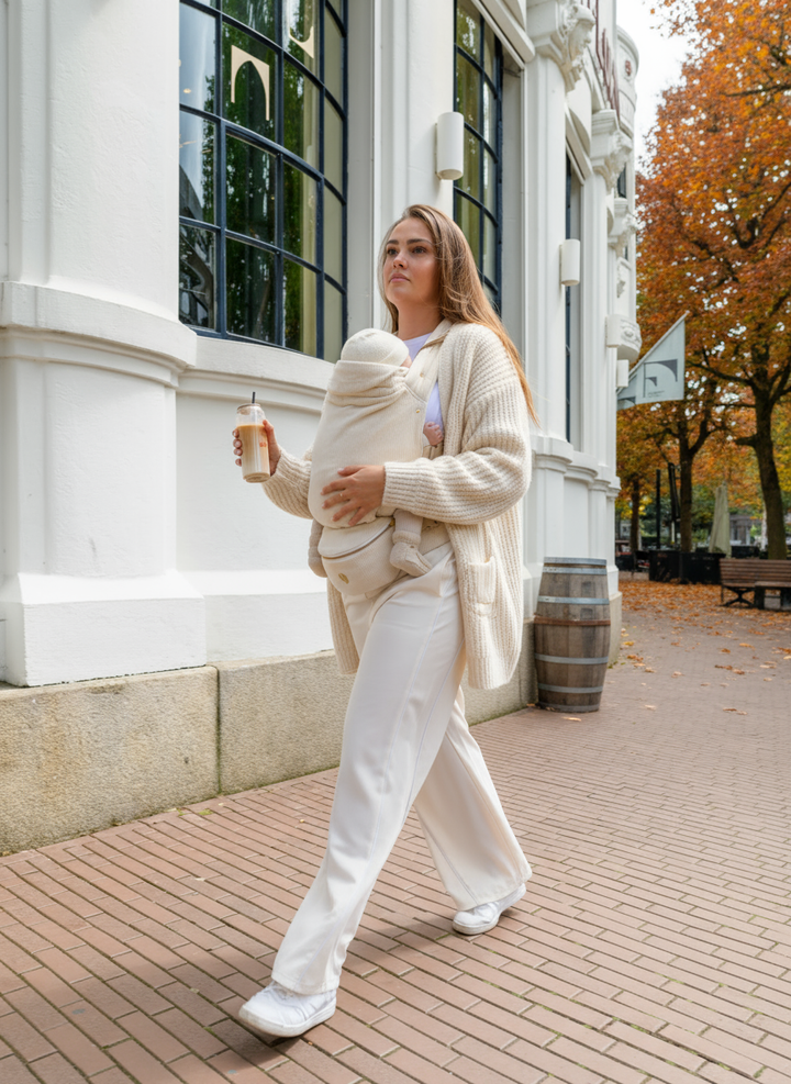 A woman walking through the city on an autumn day carrying her baby in the Babiem baby carrier Olivia. The soft off-white carrier provides comfort and closeness while matching her calm, neutral outfit and the warm seasonal atmosphere.