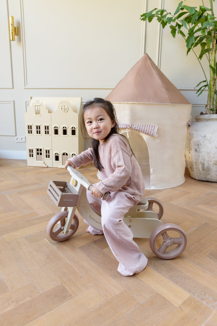 A smiling child riding the Babiem balance bike James indoors, with the Babiem dollhouse Lynn and play tent Alice visible in the background.