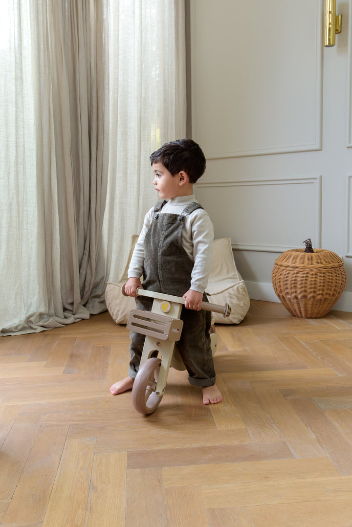 A child standing indoors while holding the Babiem balance bike James by the handlebars on a wooden floor.