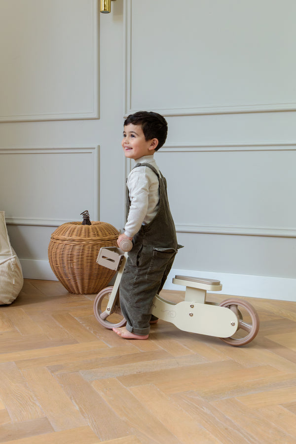 A smiling child holding and walking the Babiem balance bike James indoors, standing on a wooden floor.