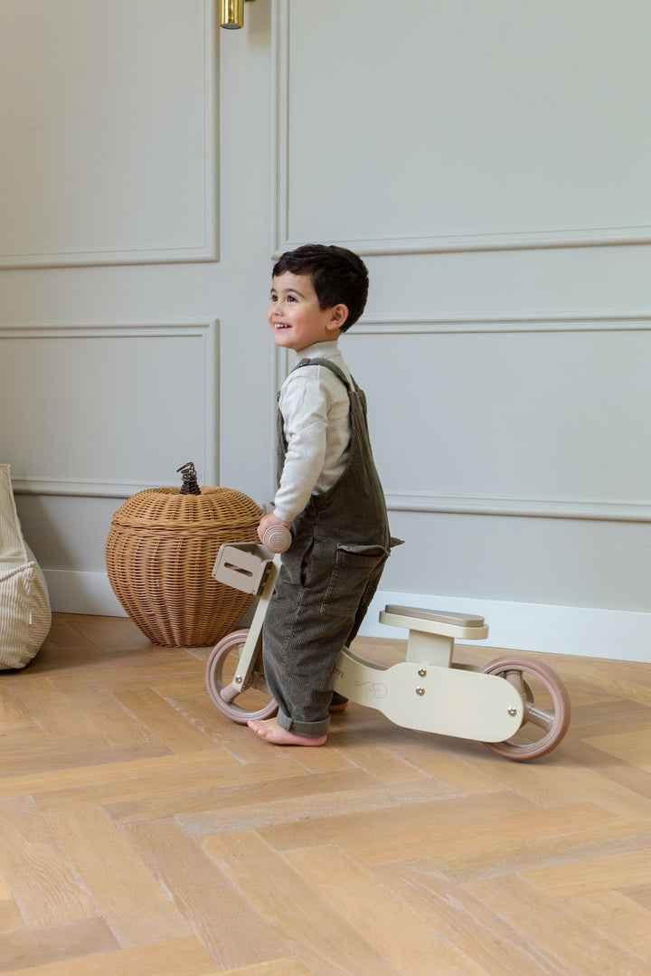 A smiling child holding and walking the Babiem balance bike James indoors, standing on a wooden floor.