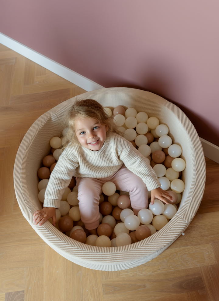 A smiling child sitting in the Babiem ball pit filled with soft-toned balls in cream, beige, and taupe. The warm pink wall and natural light create a calm, inviting atmosphere.