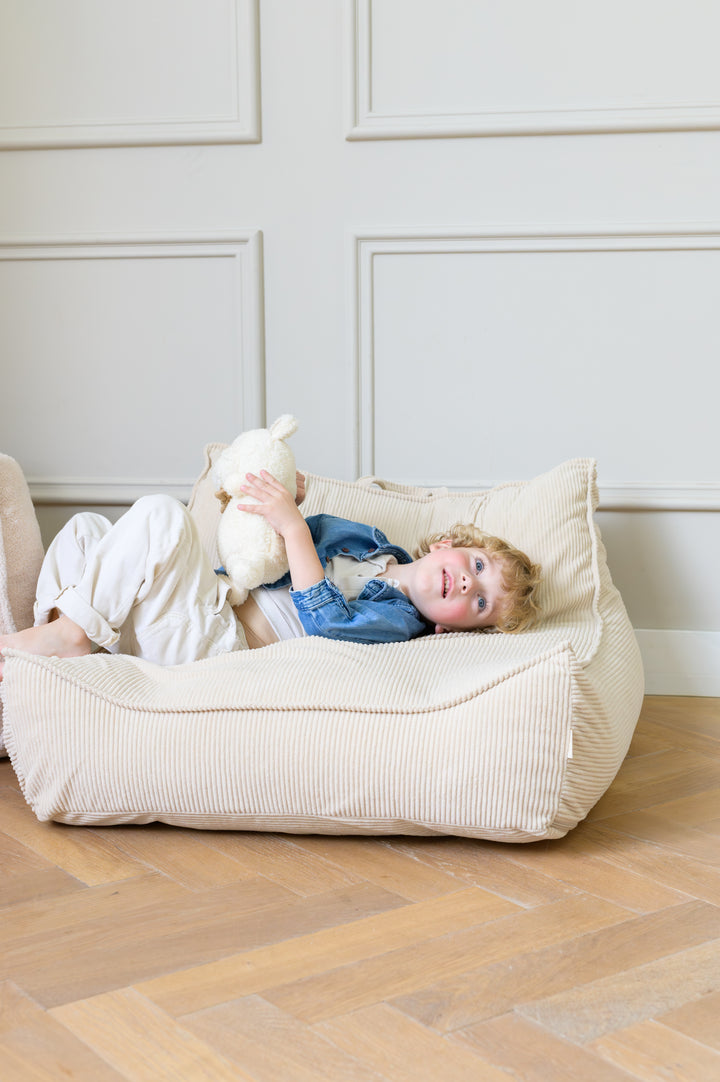 Child lying back on the Babiem beanbag Bruno in a beige fabric holding a soft toy and smiling while relaxing 