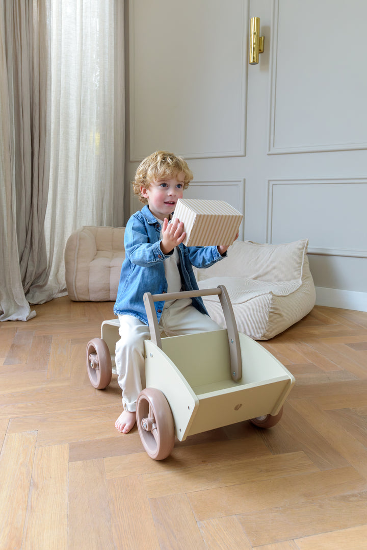 A child sitting on the Babiem cargo bike Moos while placing a striped gift box into the cargo compartment in a cozy room.