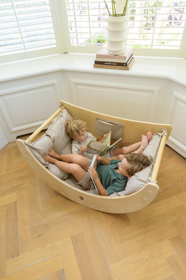 Two young children lie comfortably with books inside the Babiem climbing arch Ben, which has a cushion with soft fabric, creating a cozy and versatile play space.