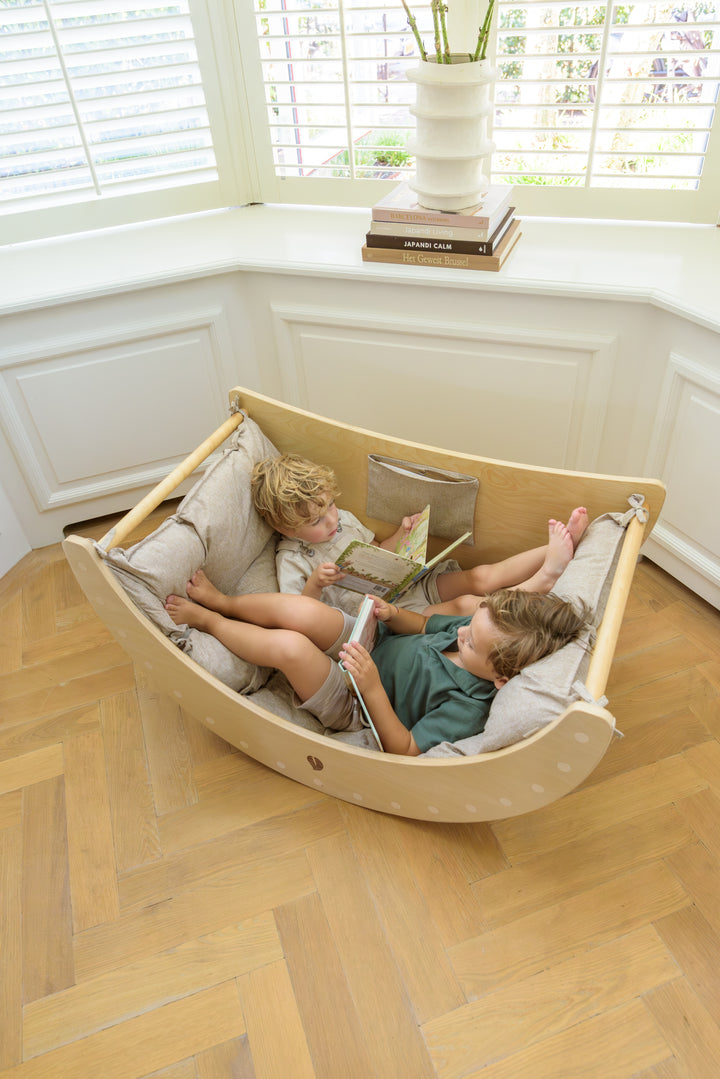 Two young children lie comfortably with books inside the Babiem climbing arch Ben, which has a cushion with soft fabric, creating a cozy and versatile play space.