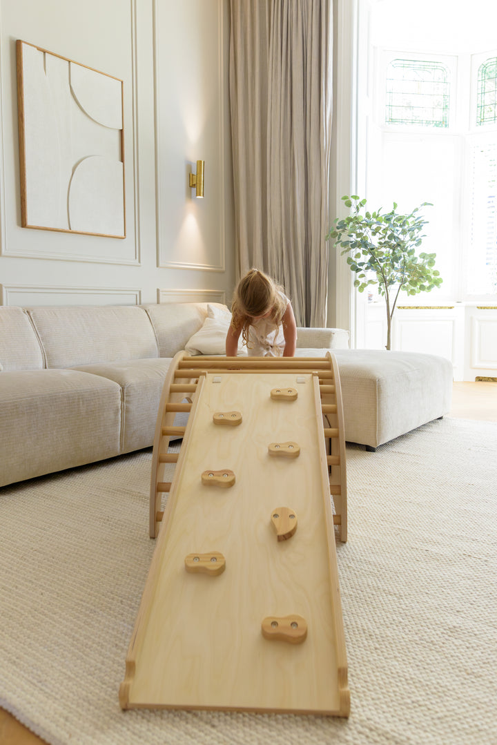 A young child in a light dress climbing up the wooden Babiem climbing arch Ben with an attached climbing ramp, placed in a cozy living room with a sofa and wall art.