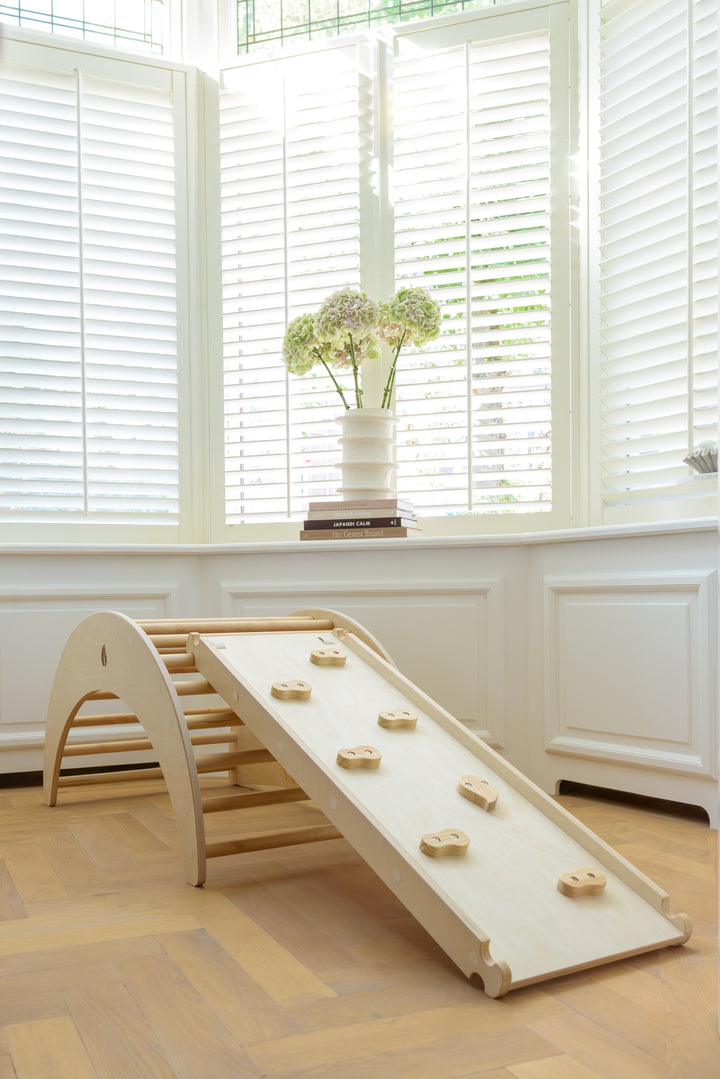 A wooden Babiem climbing arch styled as a climbing toy with a ramp and climbing holds, in a sunlit interior with white shutters and wooden floor.