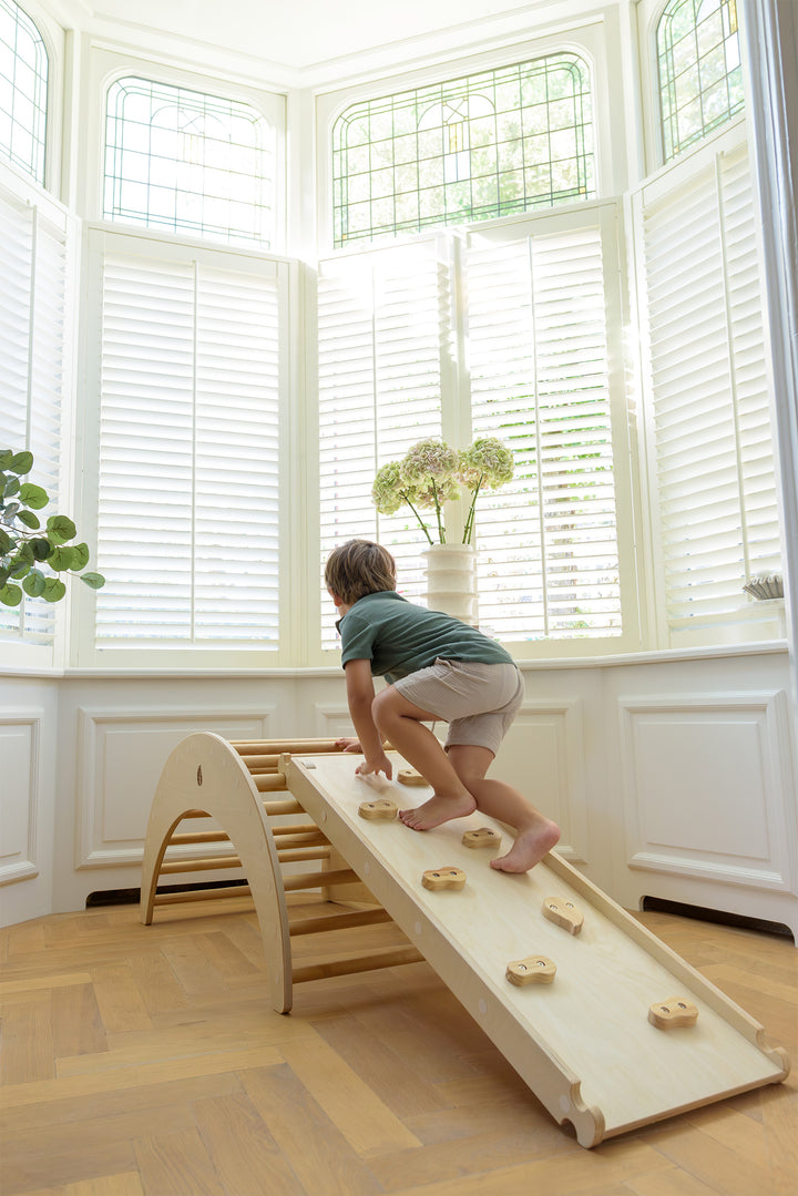 A boy in a green shirt and shorts climbing barefoot on the Babiem climbing ramp that's attached to the climbing arch, in a bright room with large windows and flowers on the sill.