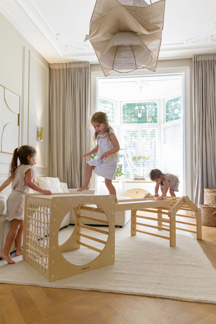 Three young children playing on the complete Babiem climbing set Ben, featuring a wooden climbing cube with rope net, a climbing house, and a ramp, placed in a bright living room with natural light.