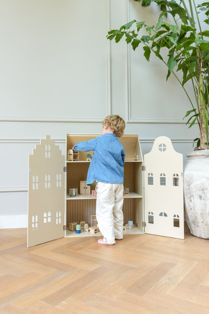 A child standing in front of the open Babiem dollhouse Lynn, arranging the miniature furniture inside a bright room with wooden flooring.