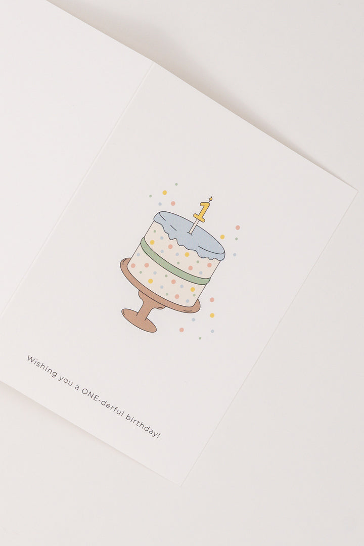 A white Babiem greeting card featuring a colorful birthday cake with a number “1” candle on top and confetti around it. The card includes the text “Wishing you a ONE-derful birthday!” printed below the illustration.