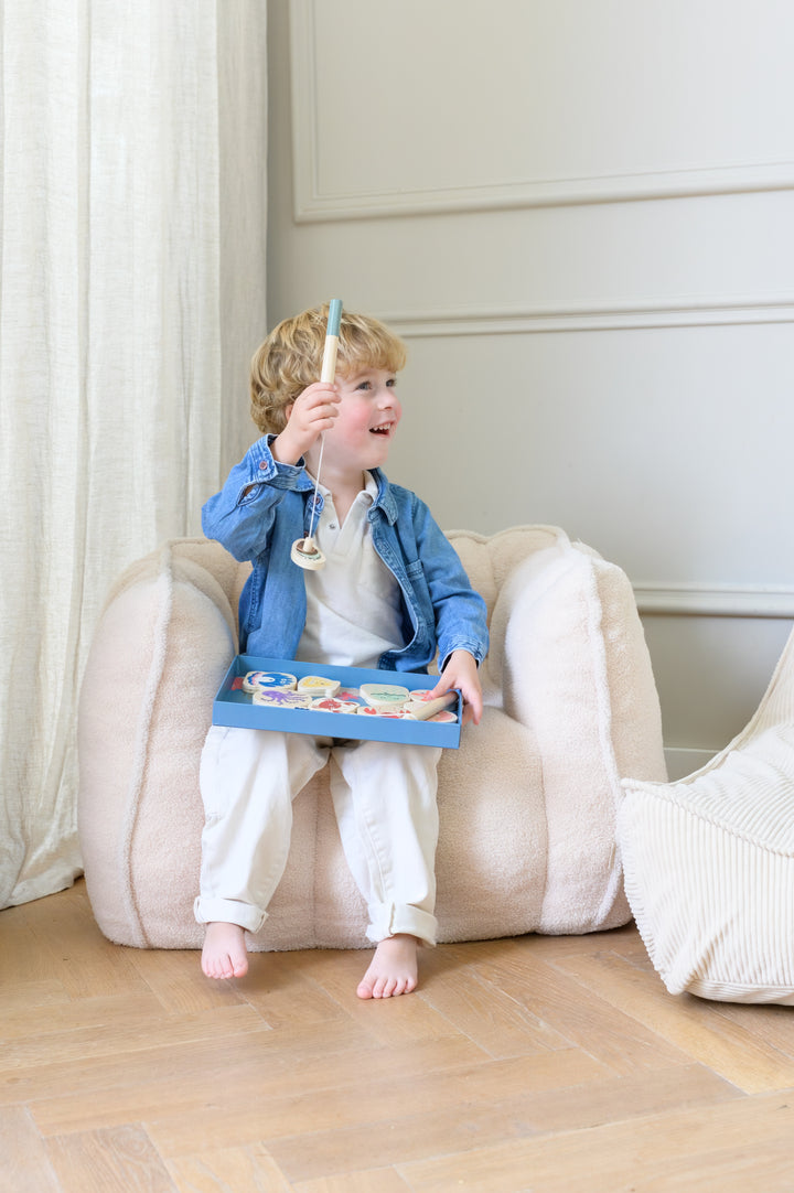 Child sitting on the Babiem kids chair Ella in a light beige fabric holding the Babiem fishing game Emma and smiling in a bright room