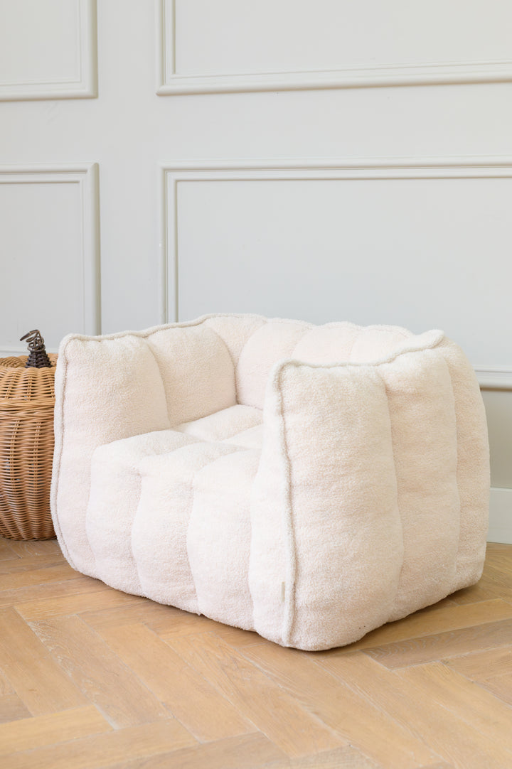 A soft, cream-colored kids chair Ella from Babiem placed on a wooden floor beside a wicker basket. The plush texture and gentle tones create a warm, inviting atmosphere perfect for a child’s room or play corner.