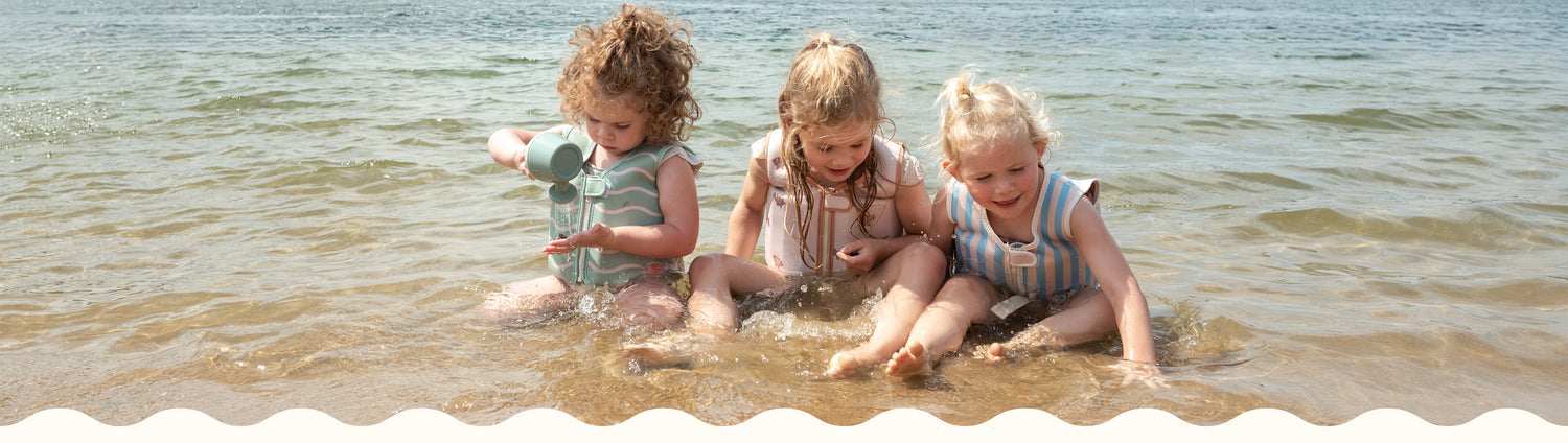 Three young children wearing Babiem swim vests sit in shallow water at the beach, playing and splashing together. Each child wears a different design: soft green waves, pink with sea stars, and blue stripes. The warm sunlight and gentle waves create a playful and carefree summer vibe, perfect for a website header.