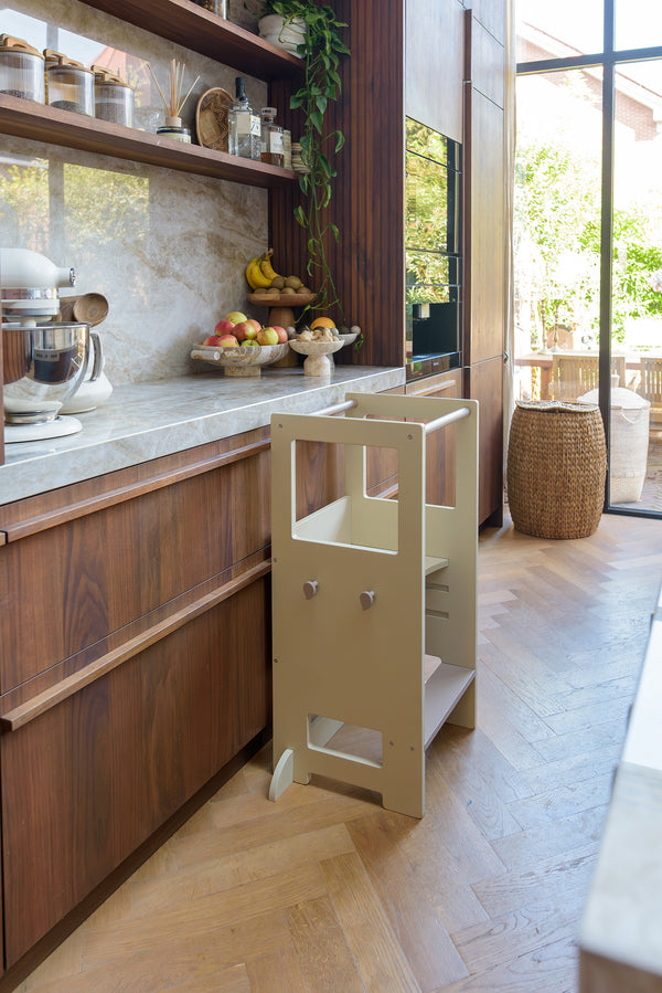 The Babiem learning tower Fay placed in a cozy, sunlit kitchen with wooden cabinets, marble countertops, and natural accents. Fresh fruit and kitchen essentials decorate the counter, creating an inviting space for little helpers to join in safely.