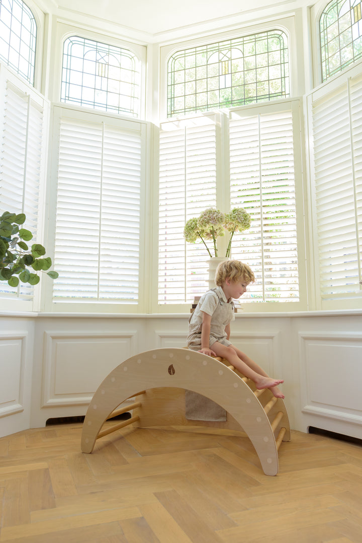 A young boy sitting on top of the wooden Babiem climbing arch, smiling in a bright room with stained glass windows and natural light.