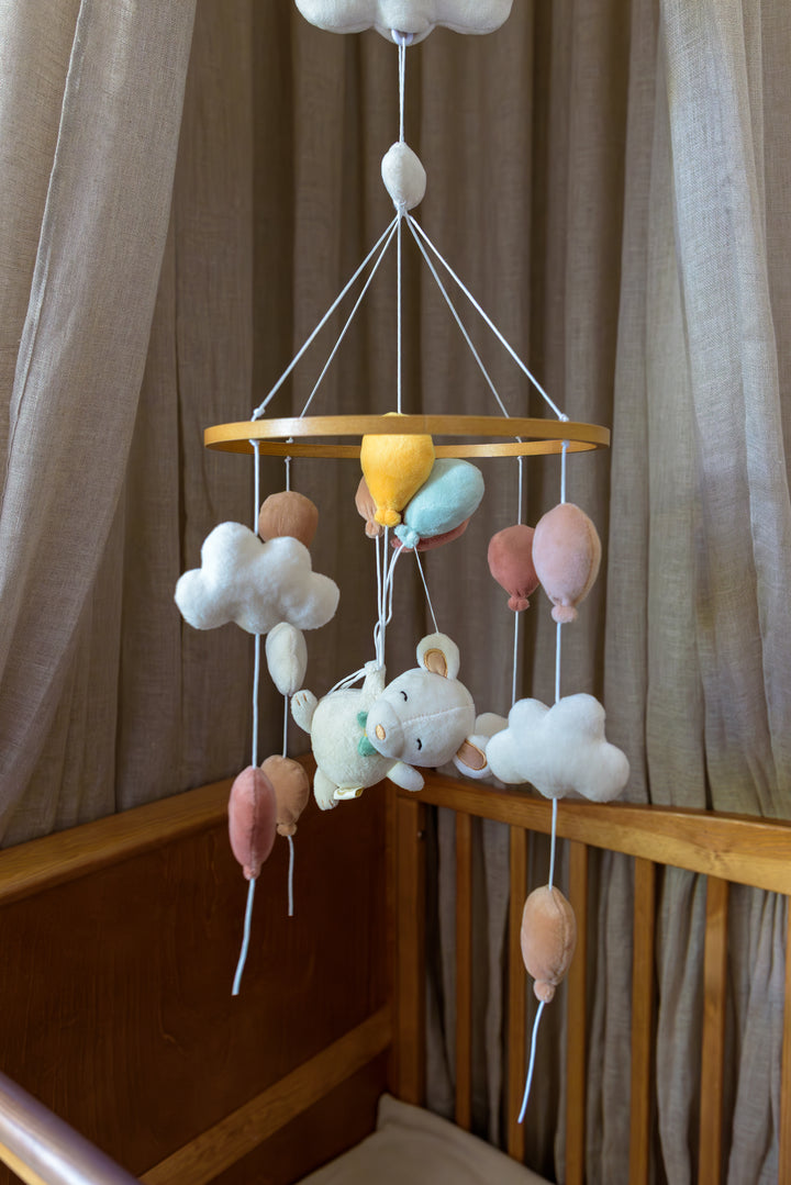 A wooden ring music mobile called Sam by Babiem, featuring a plush mouse holding colorful balloons surrounded by clouds, hanging above a crib with neutral curtains.