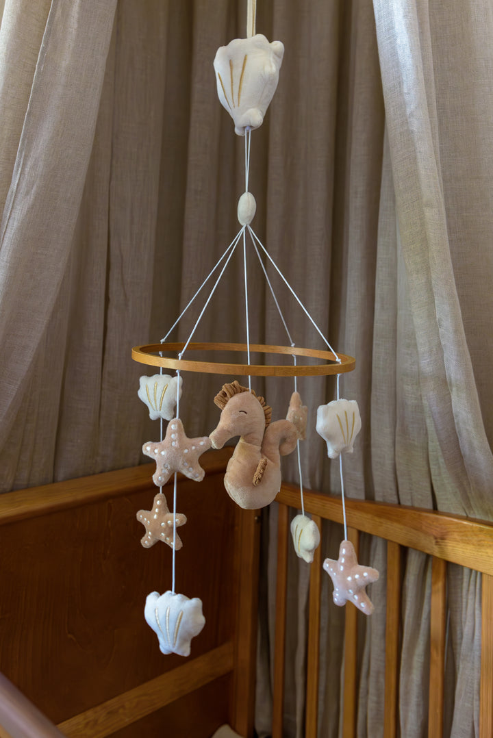 A wooden ring music mobile called Zoë by Babiem, featuring a plush seahorse surrounded by soft starfish and seashells, hanging above a crib with neutral curtains.