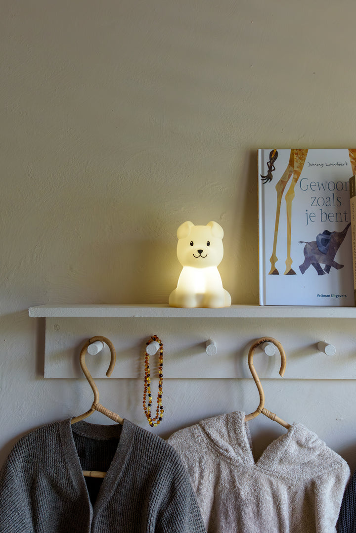 A glowing Babiem night light Bibi shaped like a dog, placed on a white shelf beside a children’s book and hanging clothes in a cozy nursery setting.