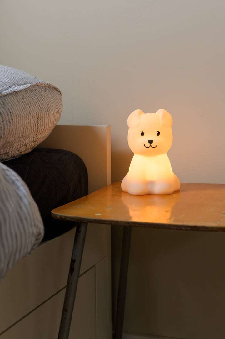 Soft glowing Babiem night light Bibi shaped like a dog, placed on a wooden bedside table next to a neatly made bed in a cozy, softly lit room.