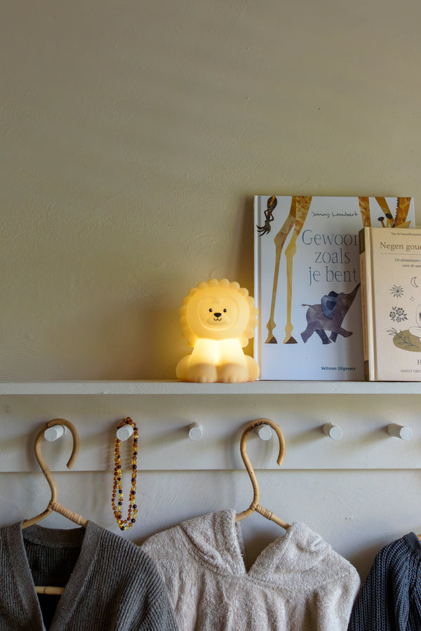 A glowing Babiem night light Lucas shaped like a lion, placed on a white shelf next to children’s books, with cozy clothes hanging below.