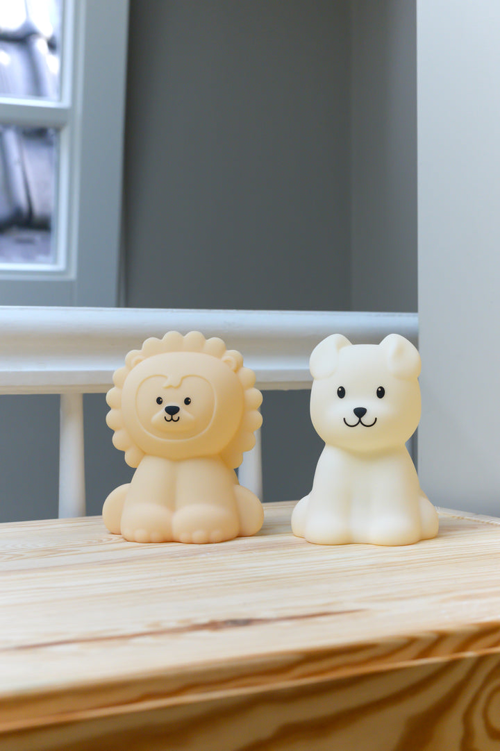 Two Babiem night lights, Bibi shaped like a dog and Lucas shaped like a lion, placed side by side on a light wooden surface near a window in a softly lit room.