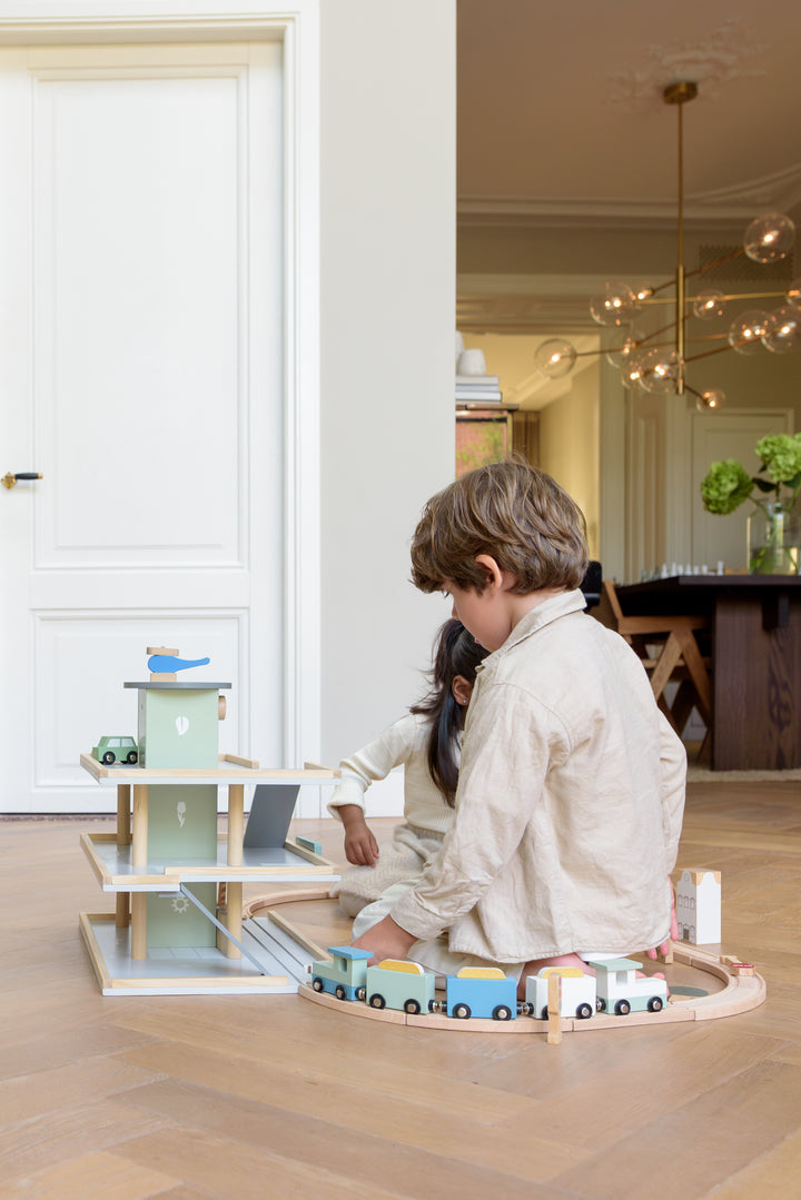 Two children playing on the floor with the Babiem play garage Noah and the wooden train set Felix, creating a playful scene in a bright living room. 