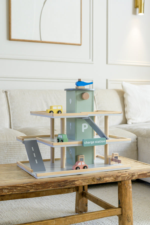 The Babiem play garage Noah shown on a wooden table, featuring multiple parking levels, toy cars, a charge station, ramps, and a blue toy helicopter on the rooftop.