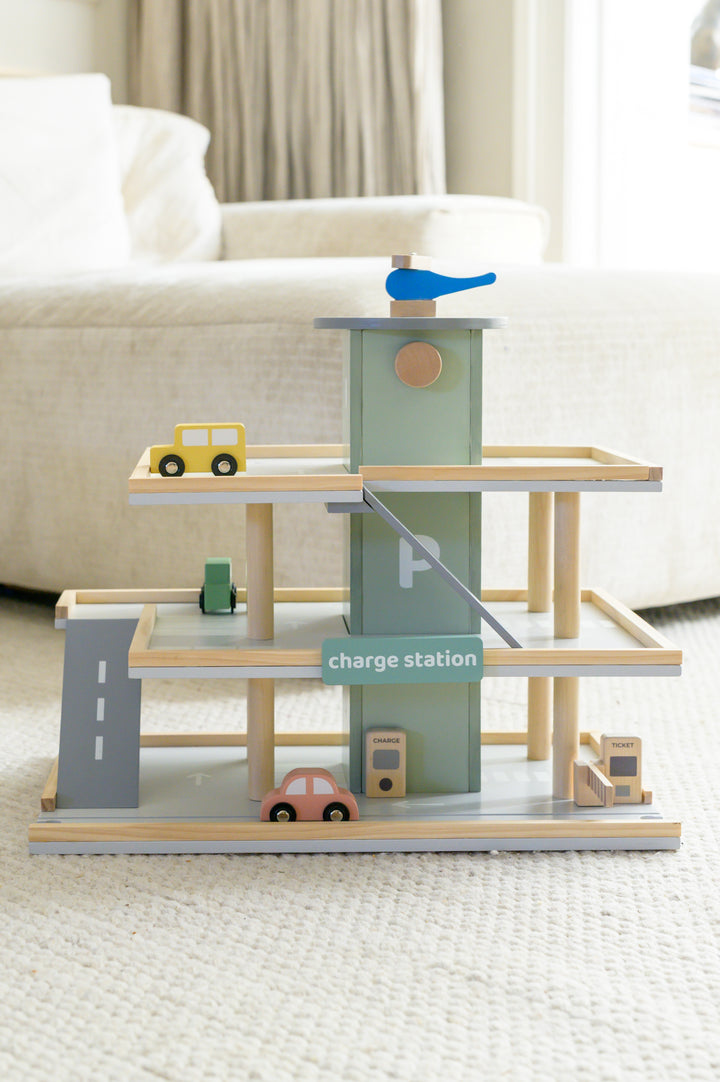 The Babiem play garage Noah displayed on a soft carpet, featuring its wooden parking levels, ramps, toy cars, a charge station, and a blue helicopter on the rooftop.