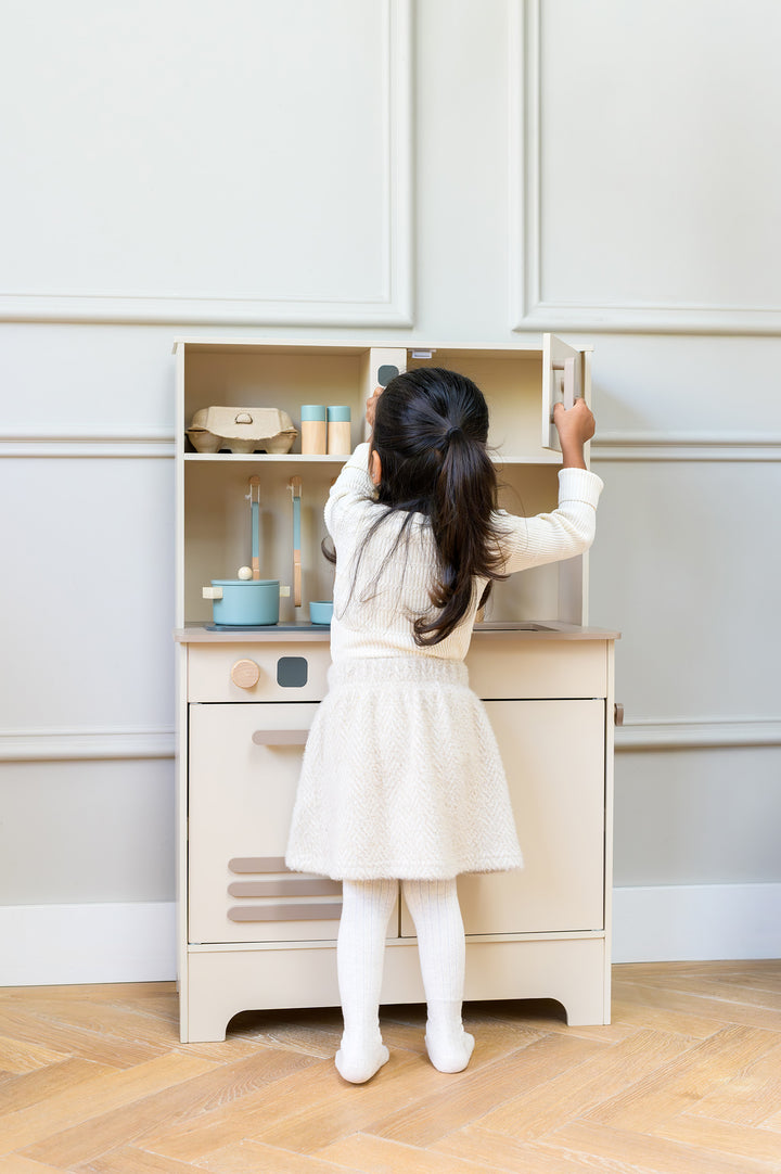 A little girl opening a cupboard of the babiem play kitchen noor, surrounded by wooden accessories in a softly lit room with light walls and wooden flooring