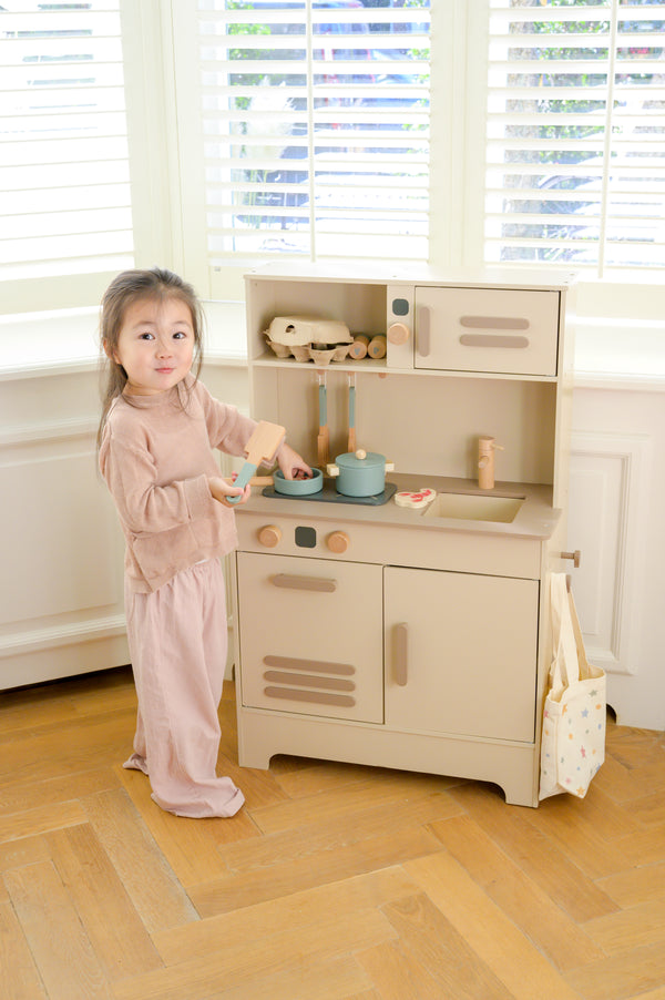 A smiling child standing at the Babiem play kitchen Noor, holding a wooden spatula while pretend-cooking with toy pots and food items.