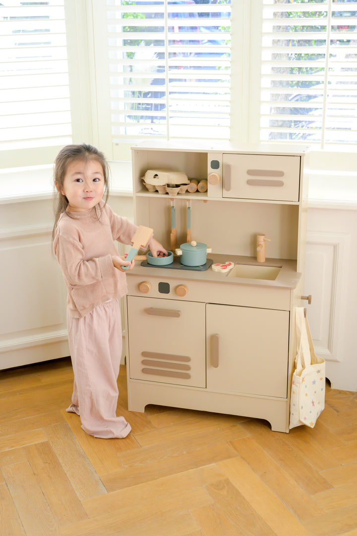 A smiling child standing at the Babiem play kitchen Noor, holding a wooden spatula while pretend-cooking with toy pots and food items.