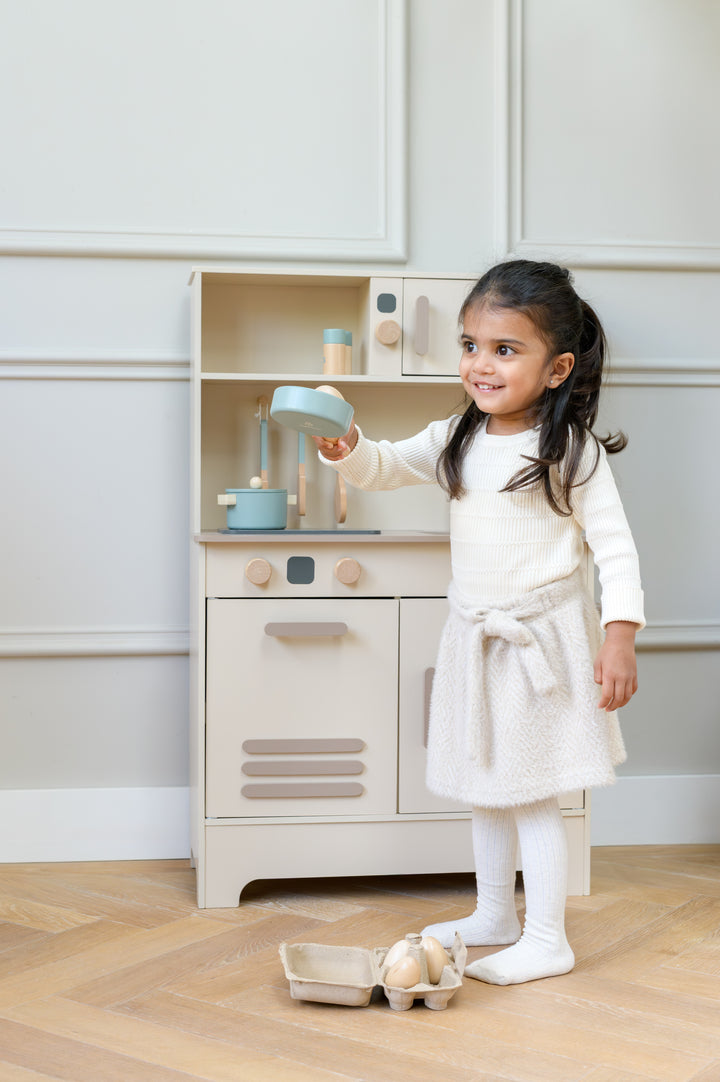 A smiling child holding a toy cooking pan while standing in front of the Babiem play kitchen Noor, with pretend eggs on the floor nearby.
