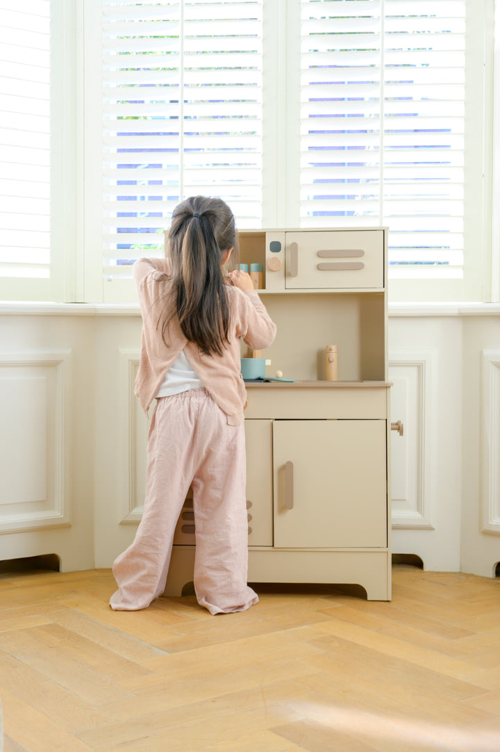 A child standing at the Babiem play kitchen Noor, pretending to prepare food in a bright room with white shutters and wooden flooring.
