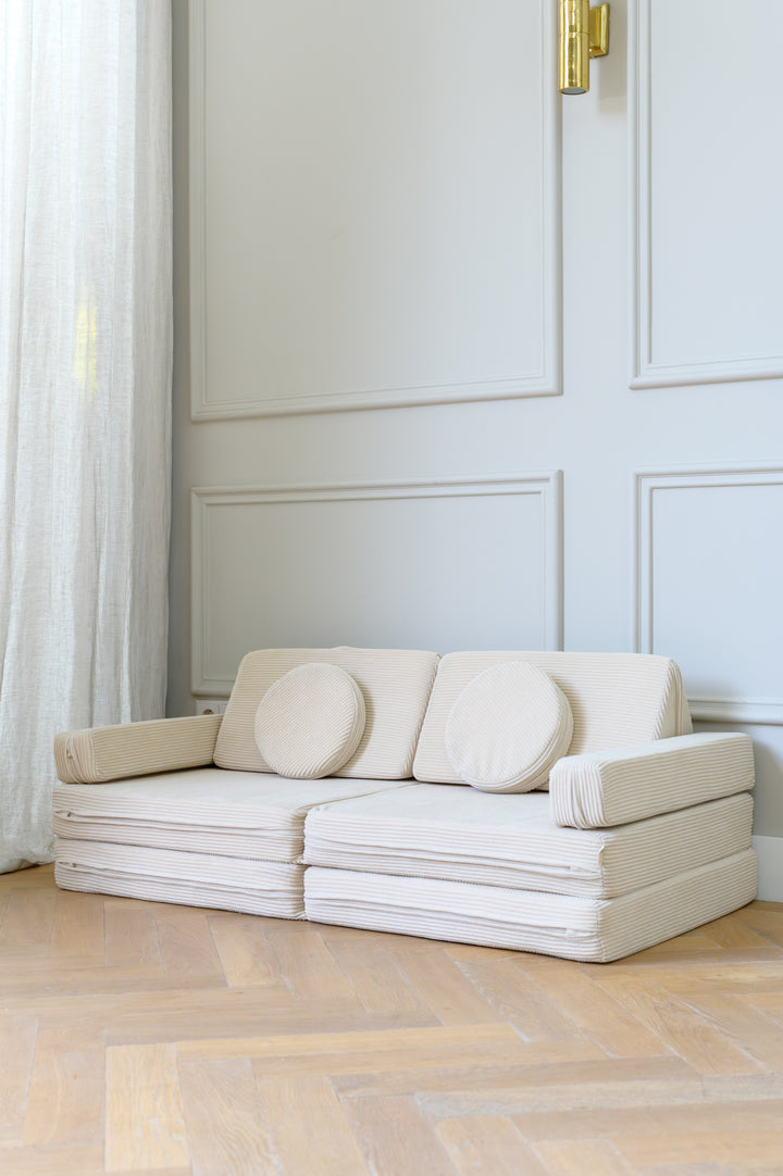 The Babiem play sofa Max set up in a bright room with soft curtains and wooden flooring, featuring layered cushions and round pillows in a neutral corduroy fabric.