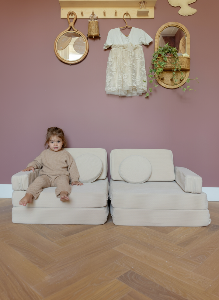 A young child sitting on the Babiem play sofa Max against a warm mauve wall decorated with natural wooden and rattan accessories. The soft off-white cushions and calm tones create a cozy, inviting atmosphere perfect for play and relaxation.