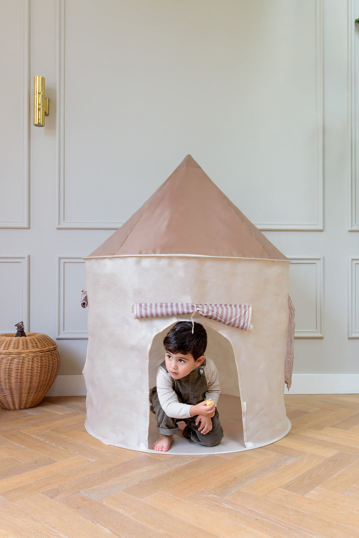 A young child plays inside the Babiem play tent Alice, a soft beige and taupe and beige-colored fabric playhouse with a conical roof, creating a cozy and imaginative space for tiny ones to explore indoors.