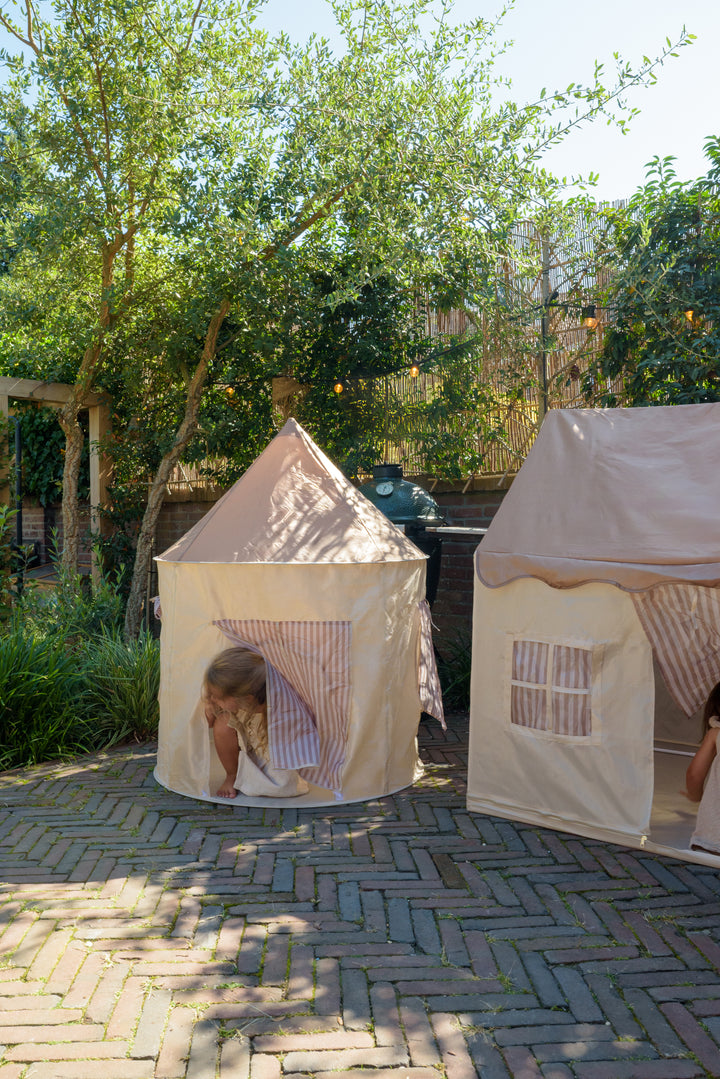 A girl peeking out of the Babiem play tent Alice and a matching fabric playhouse, placed on a brick terrace surrounded by trees and plants.