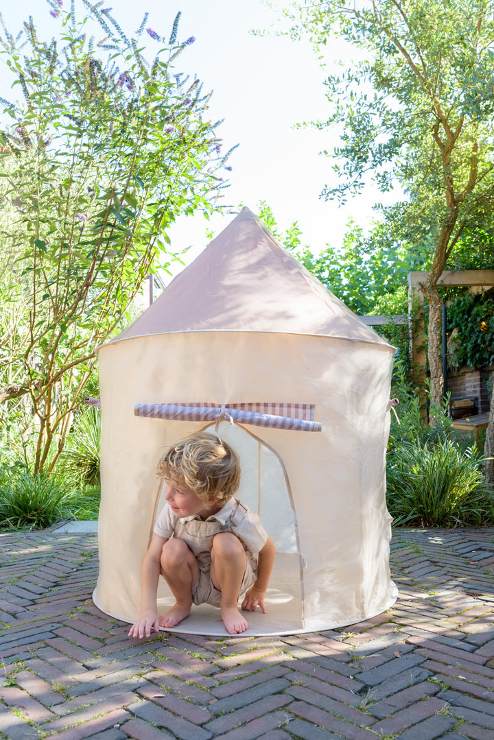 A young boy in a beige romper crouching at the entrance of the round Babiem play tent Alice, set outdoors on a sunny day with greenery around.