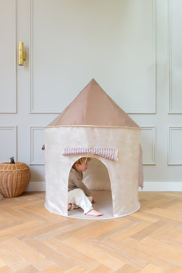 A child sitting inside the Babiem play tent Alice in a bright room with wooden flooring and a woven basket nearby.
