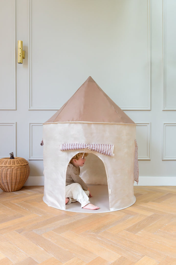 A child sitting inside the Babiem play tent Alice in a bright room with wooden flooring and a woven basket nearby.