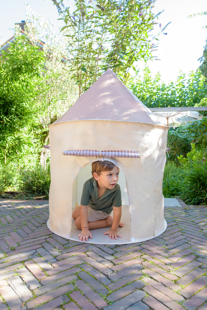 A boy in a green shirt and shorts crawling out of the Babiem play tent Alice, styled with a soft beige fabric and striped door, in a green garden.
