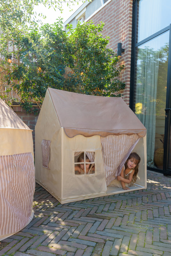 Children playing in the Babiem play tent Omar, peeking out through the striped curtains and window, placed outdoors on a brick terrace.