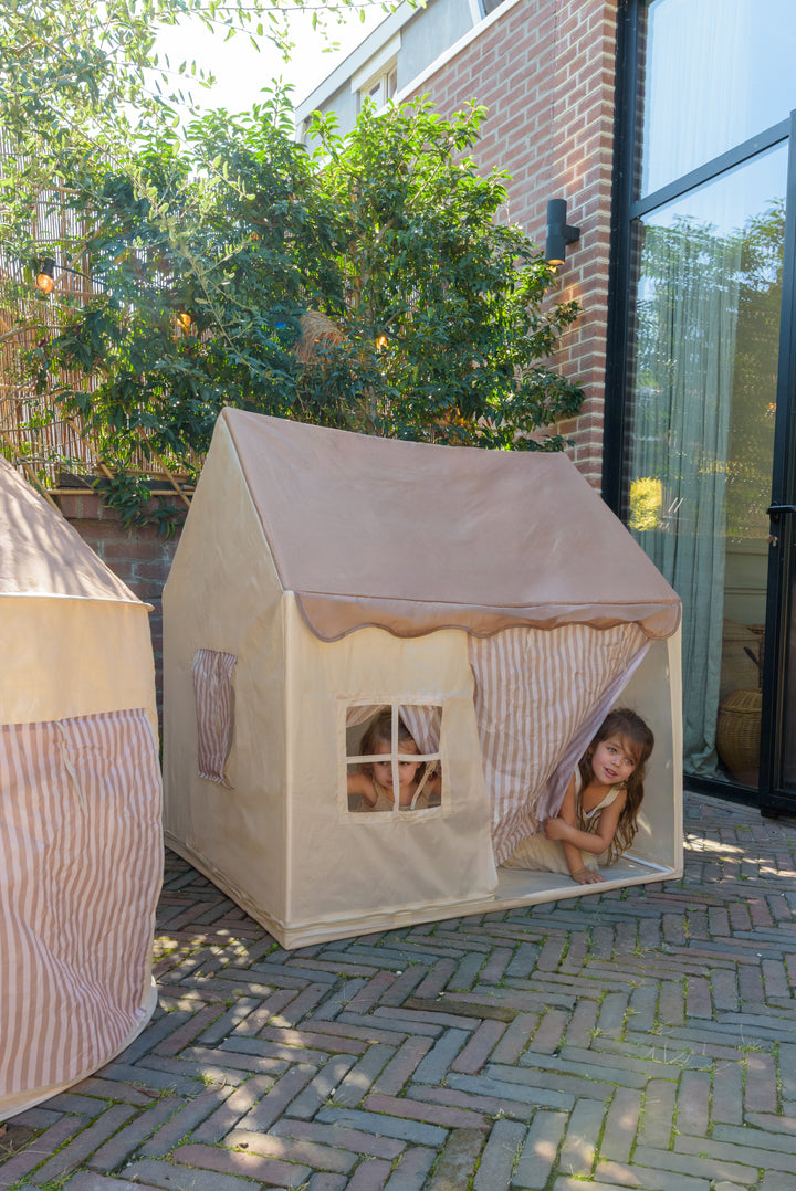 Children playing in the Babiem play tent Omar, peeking out through the striped curtains and window, placed outdoors on a brick terrace.