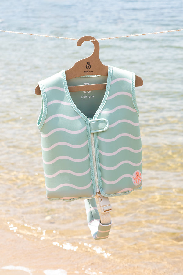 Babiem swim vest in mint green with white wave pattern and a small orange octopus motif, hanging on a branded wooden hanger against a calm seaside backdrop. The vest includes a white buckle strap visible at the bottom.